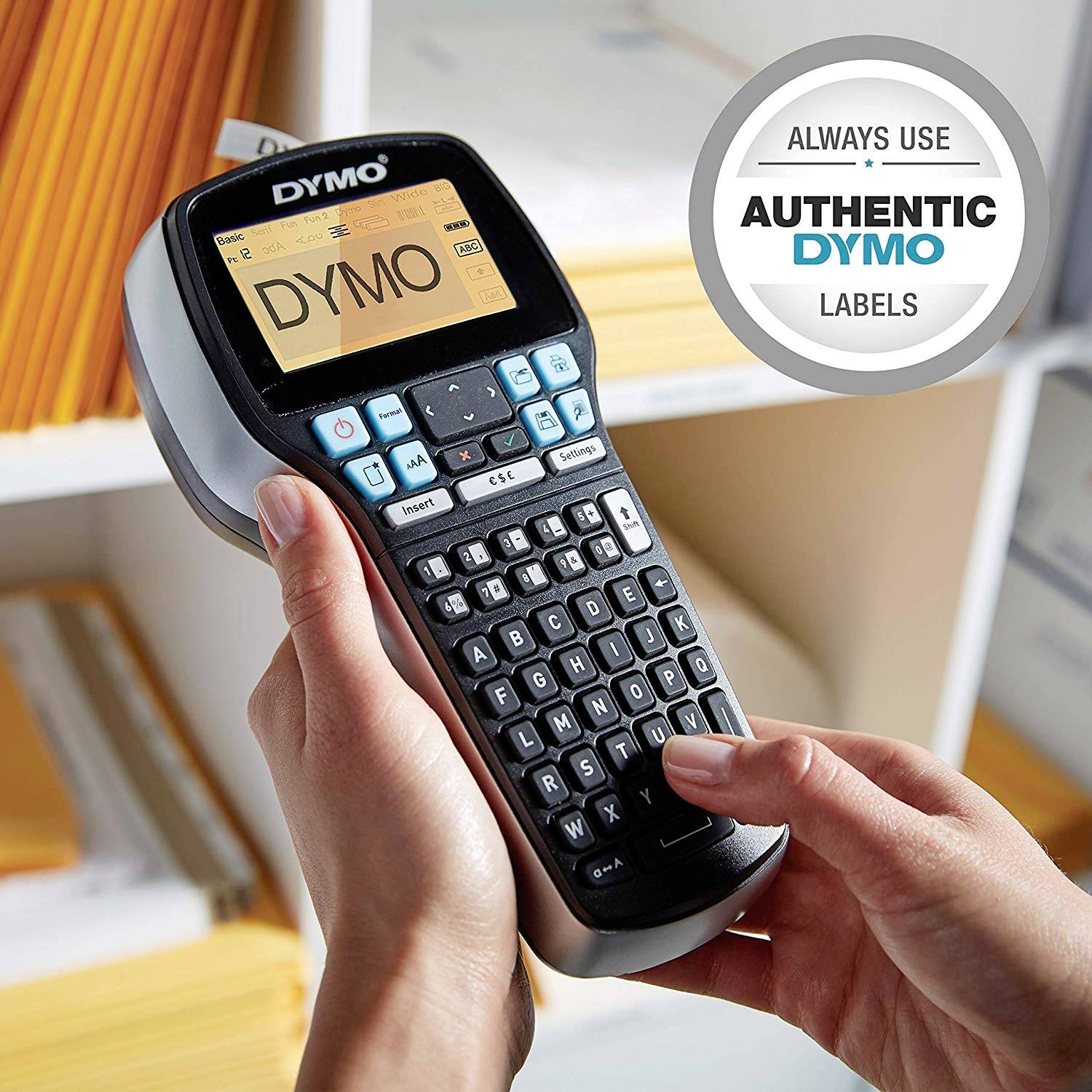 A person is holding a label printer in their hand, with 'DYMO' displayed on the screen. Bookshelves are visible in the background. A round sticker in the top right corner encourages the use of 'AUTHENTIC DYMO' labels.