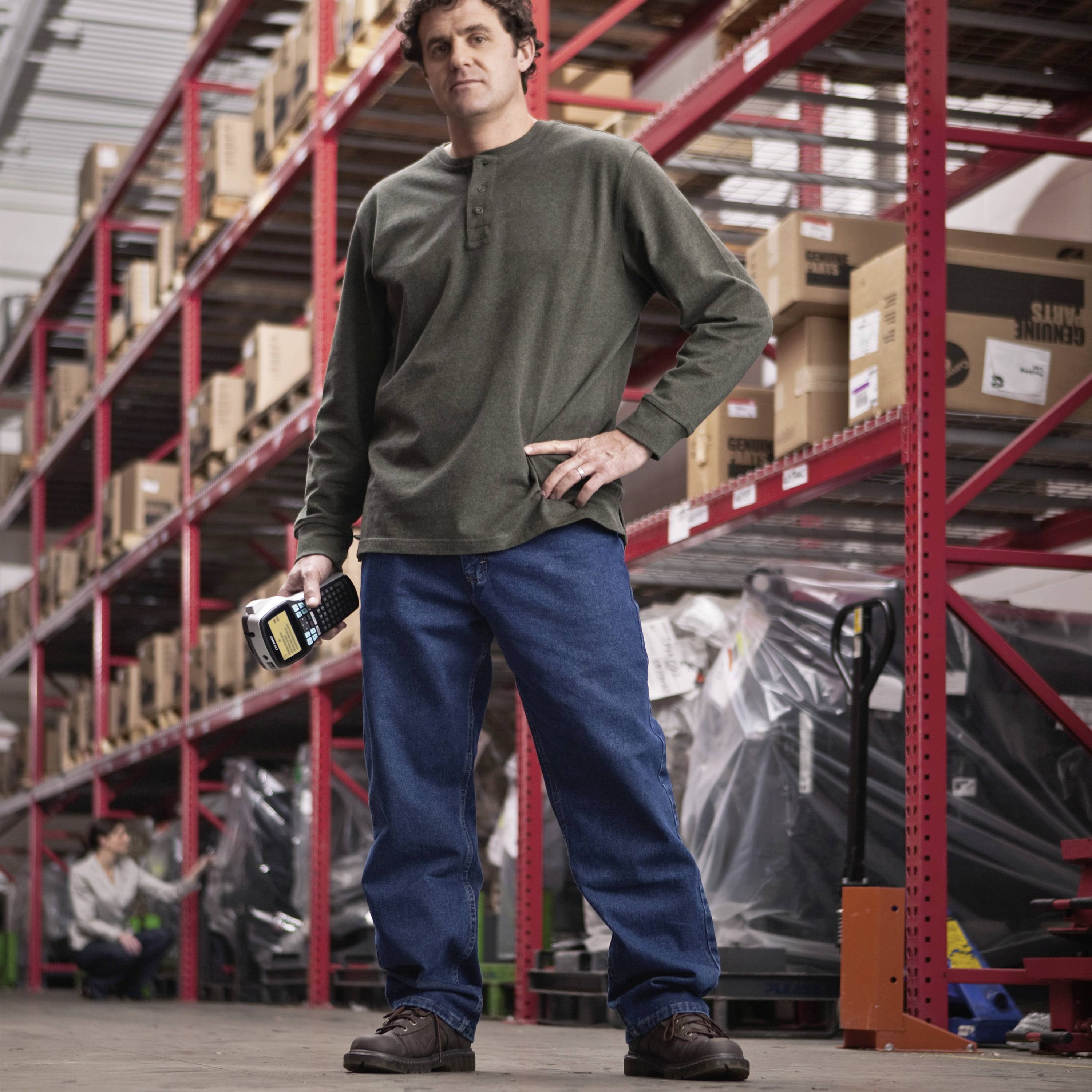 A person is standing in a warehouse with shelves full of boxes. They are holding a cardboard labelling device and wearing casual clothing.