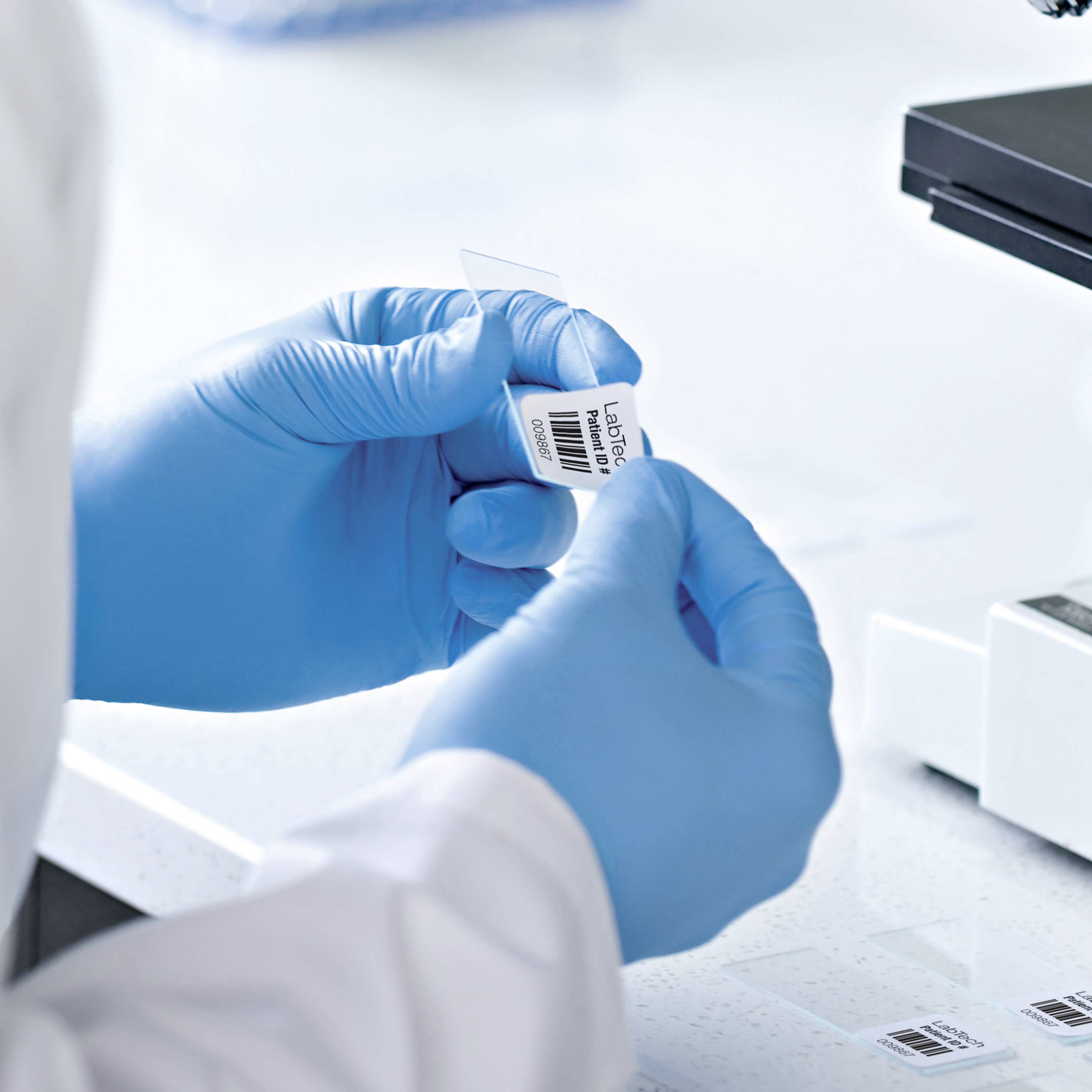 A person wearing blue gloves is holding a test tube and a label. A scanner is lying on a table nearby.