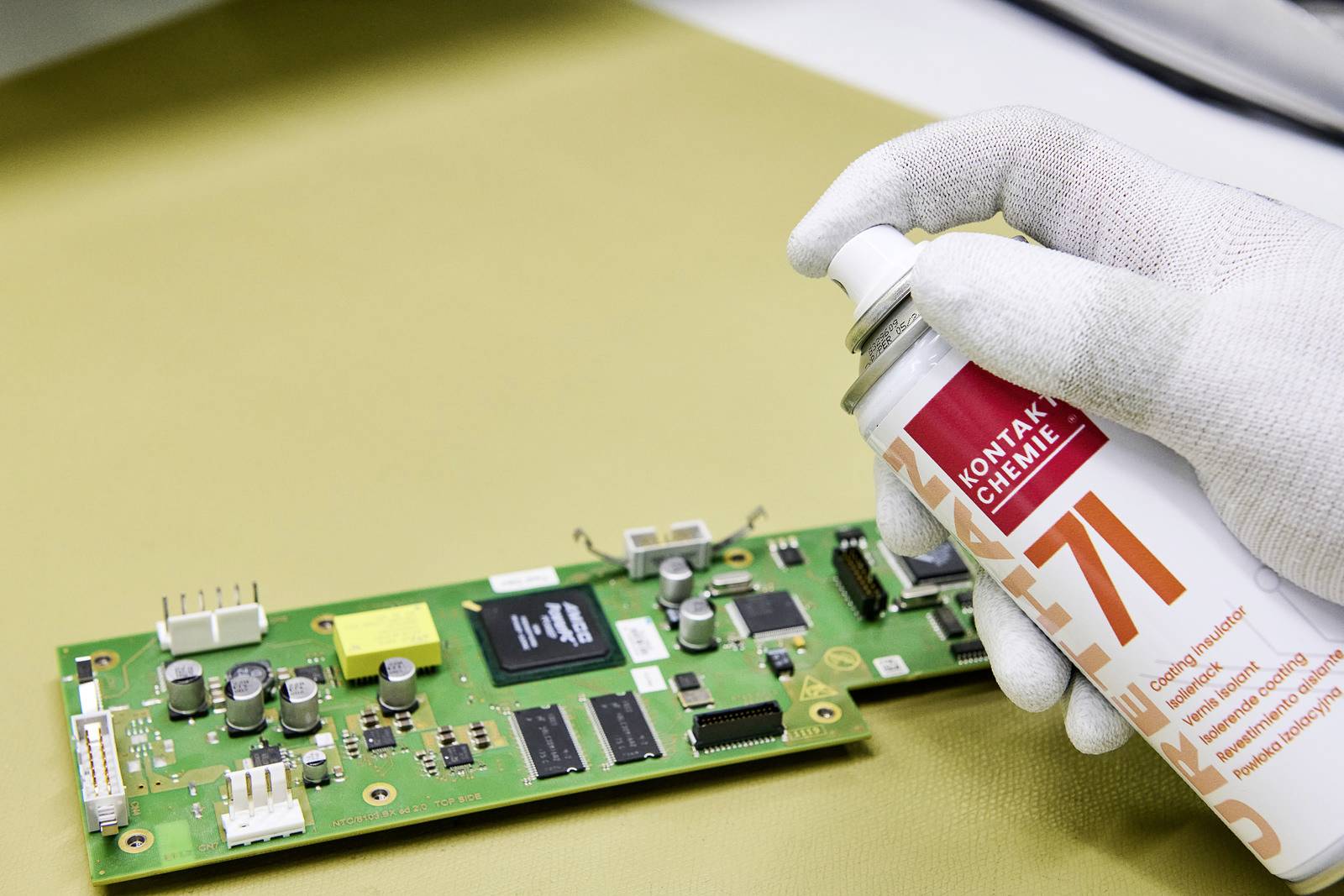 A gloved hand sprays a protective agent from a can onto a green circuit board with electronic components.