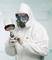 A person in protective clothing and a respiratory mask is spraying a substance, possibly in a laboratory or workshop environment.