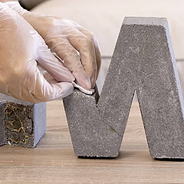 A gloved hand carefully shapes a gray, concrete-like sculpture, smoothing its surface with a tool, implying a focus on craftsmanship.