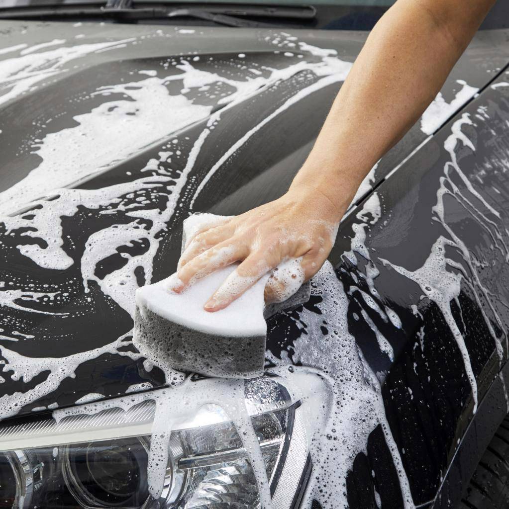 A person is washing a car by hand, rubbing soap with a sponge onto the bonnet. The headlight is visible.