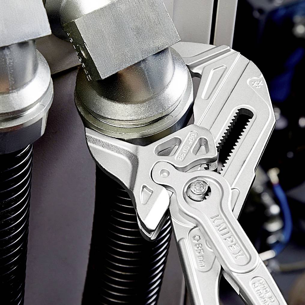 A pipe wrench grips a metal pipe next to another pipe. Both pipes are connected to fittings. Tool application in focus.