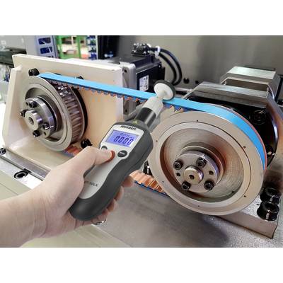A hand holds a digital device measuring the tension of a blue-grey belt between two large gears.