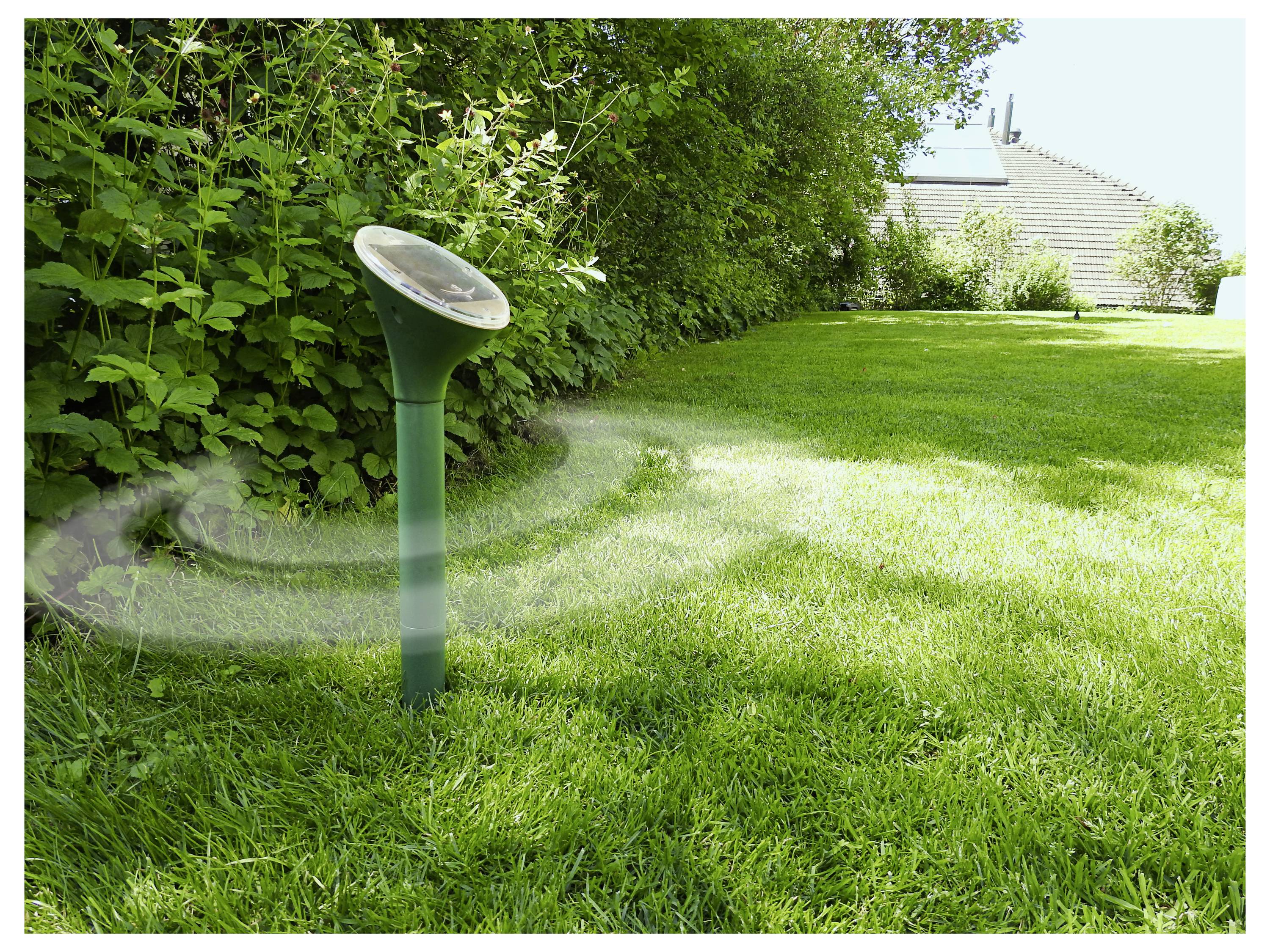 A green lawn with a solar-powered device emitting sound waves to deter moles, set against a backdrop of trees and a house.
