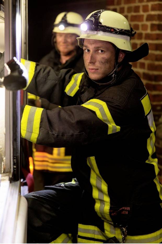 Two firefighters in protective clothing and helmets are working intently inside a building. They are wearing head torches and have serious expressions.