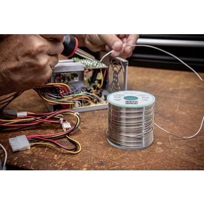 A person is soldering cables onto an electronic device. A soldering wire is in the foreground on a wooden table.