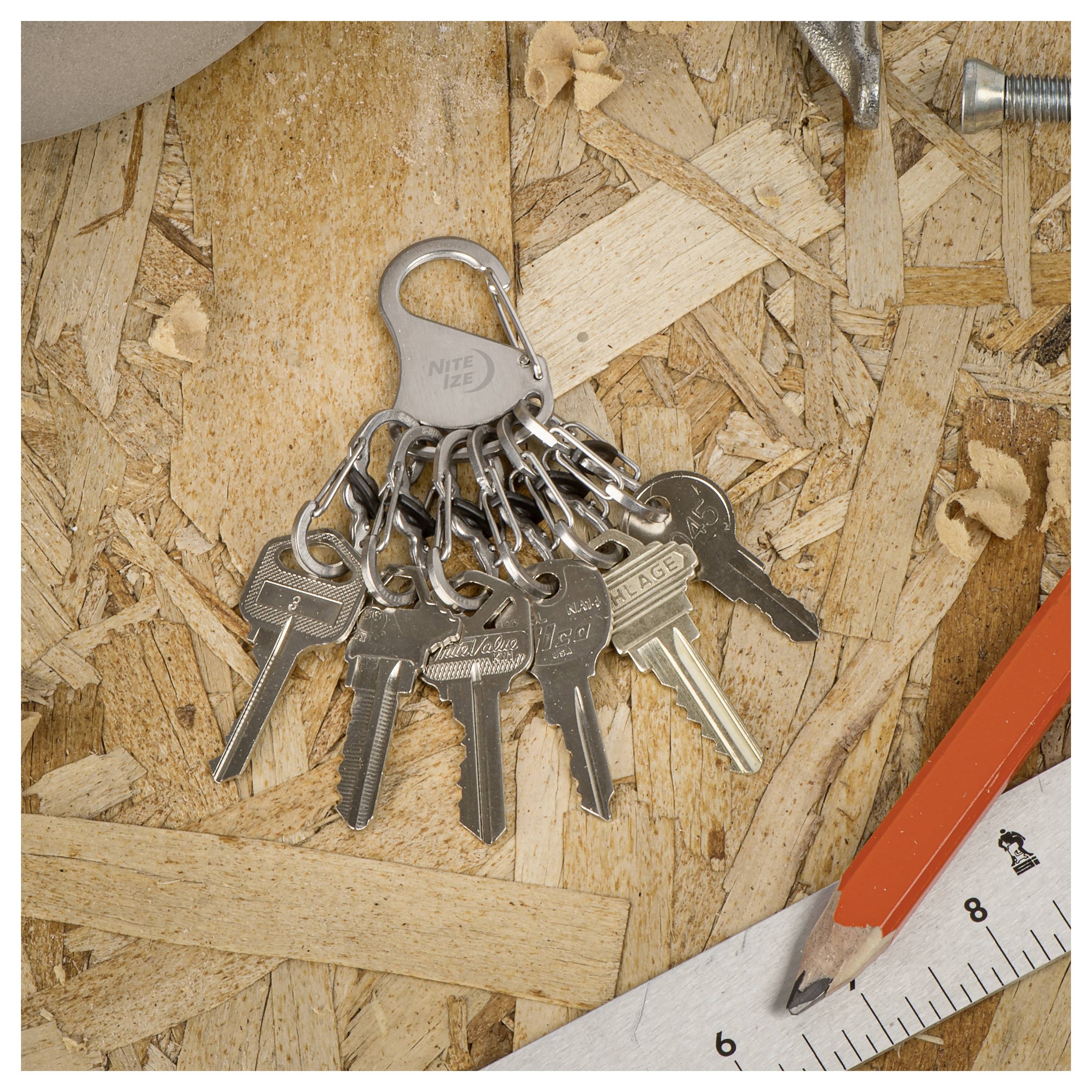 A set of nine metal keys on a chain sits on a wooden surface, surrounded by a steel hook, silver screw, red pencil, and a white ruler.