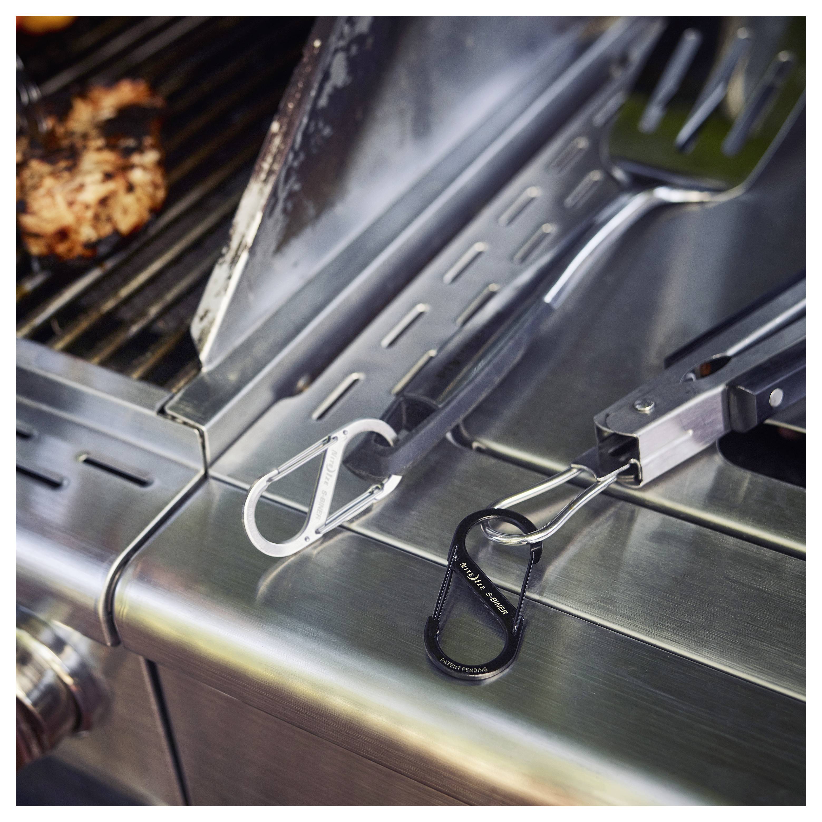 A metal grill with meat cooking; two pairs of tongs with carabiner clips resting on the grill's side shelf.