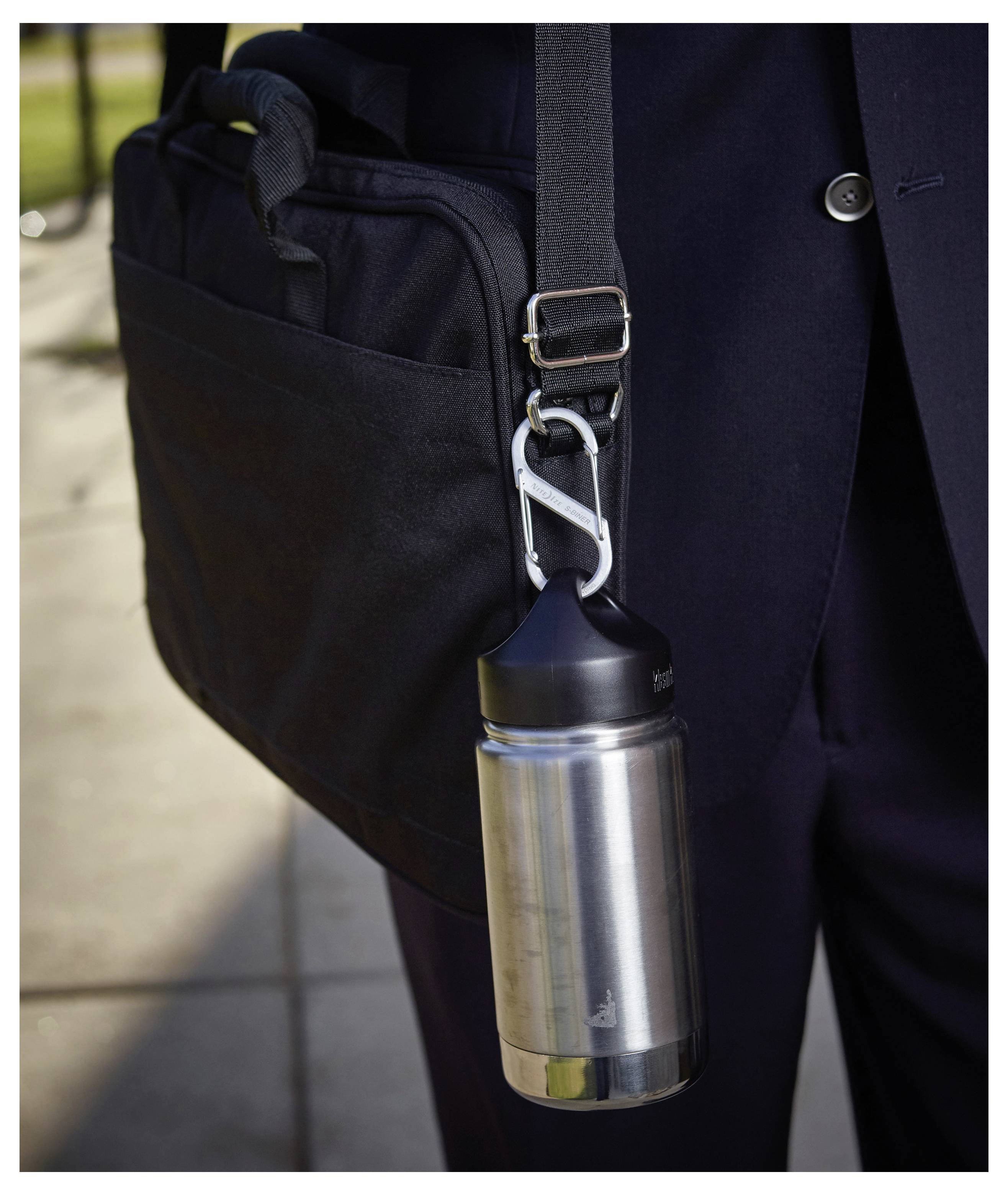 A stainless steel water bottle attached to a black shoulder bag with a carabiner, carried by a person in a dark suit outdoors.