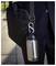 A stainless steel water bottle attached to a black shoulder bag with a carabiner, carried by a person in a dark suit outdoors.