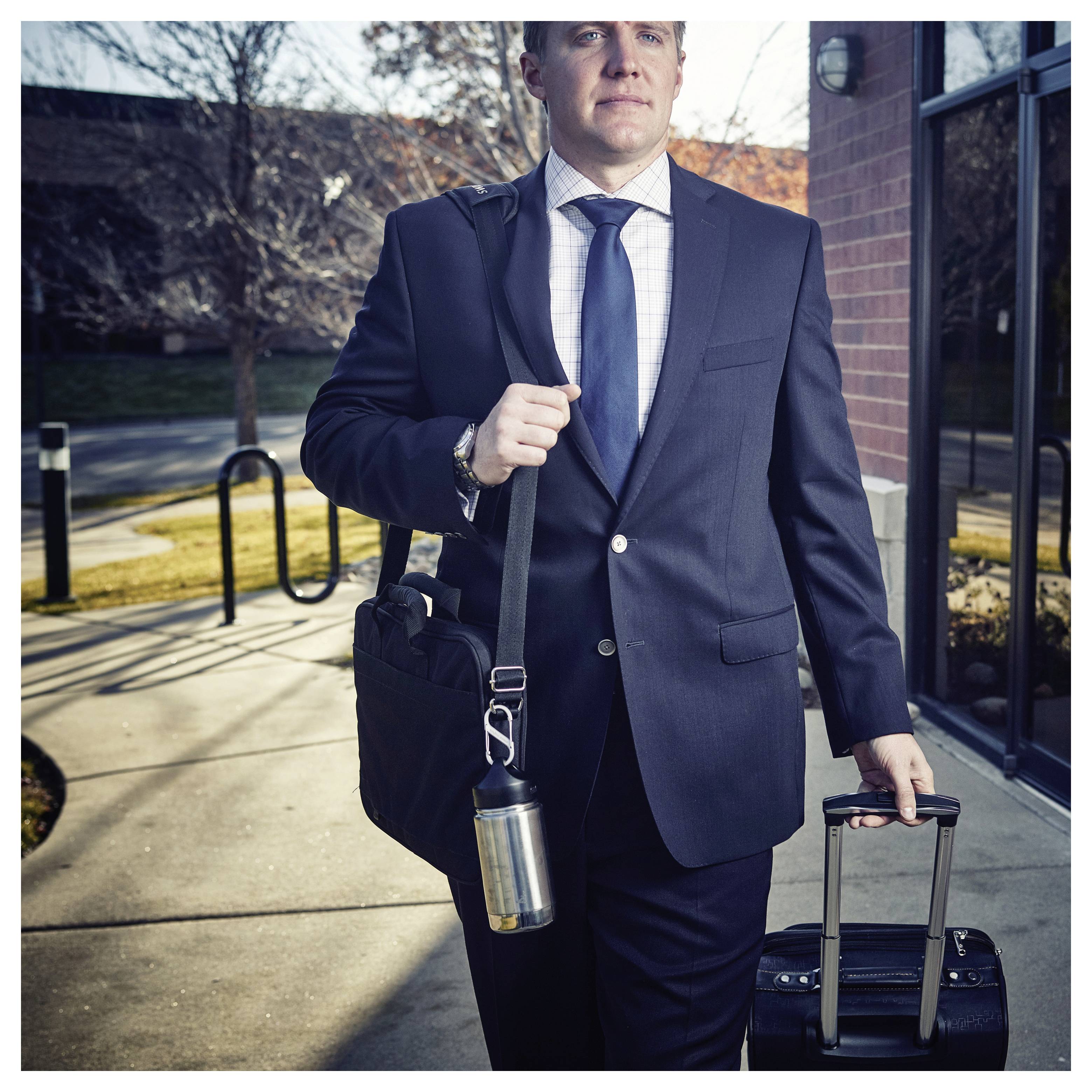 Man in a suit walking outside, holding a briefcase and pulling a rolling suitcase. He appears focused and professional.