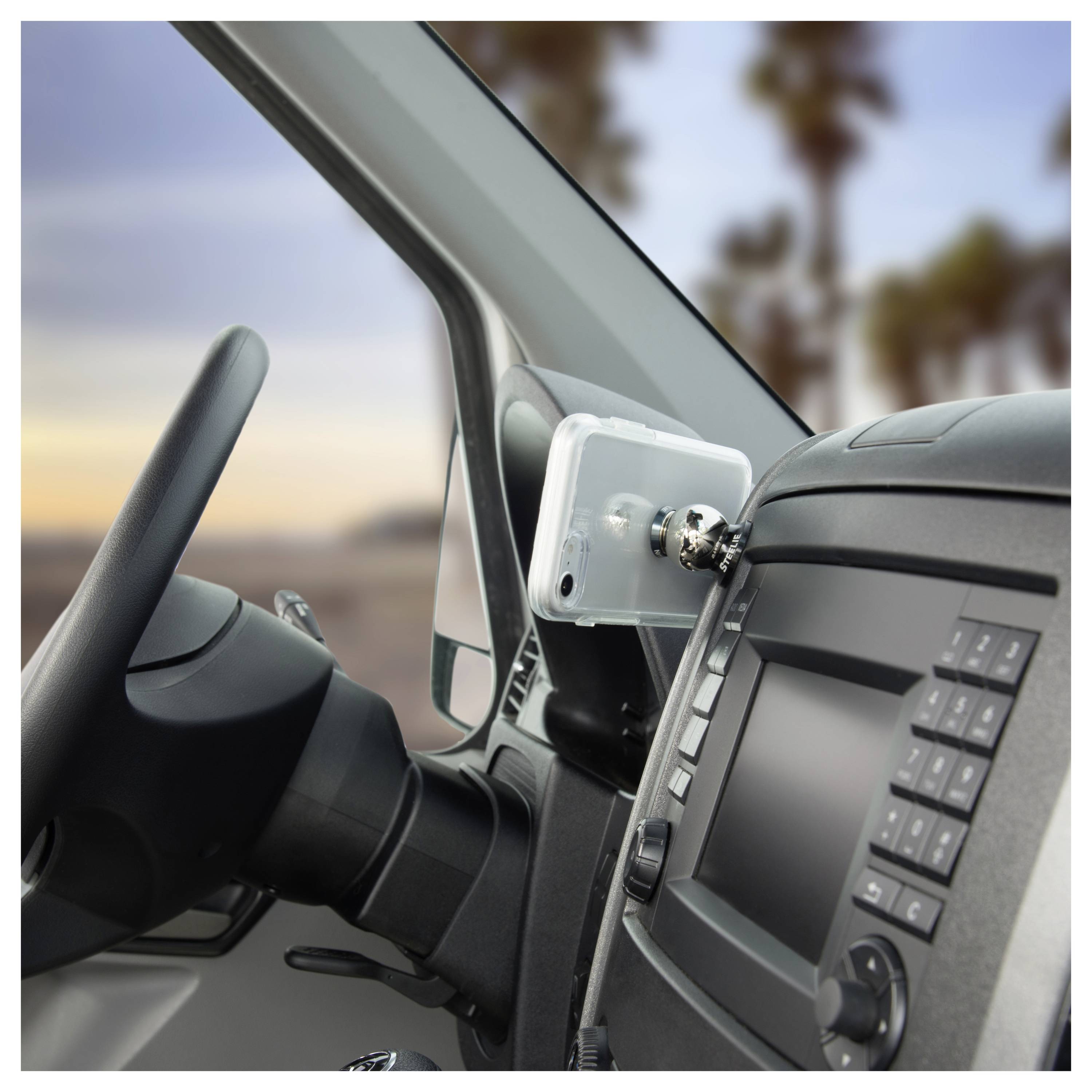 Phone mounted on a car dashboard near steering wheel, next to a control panel. Blurred background shows a sunset and palm trees.