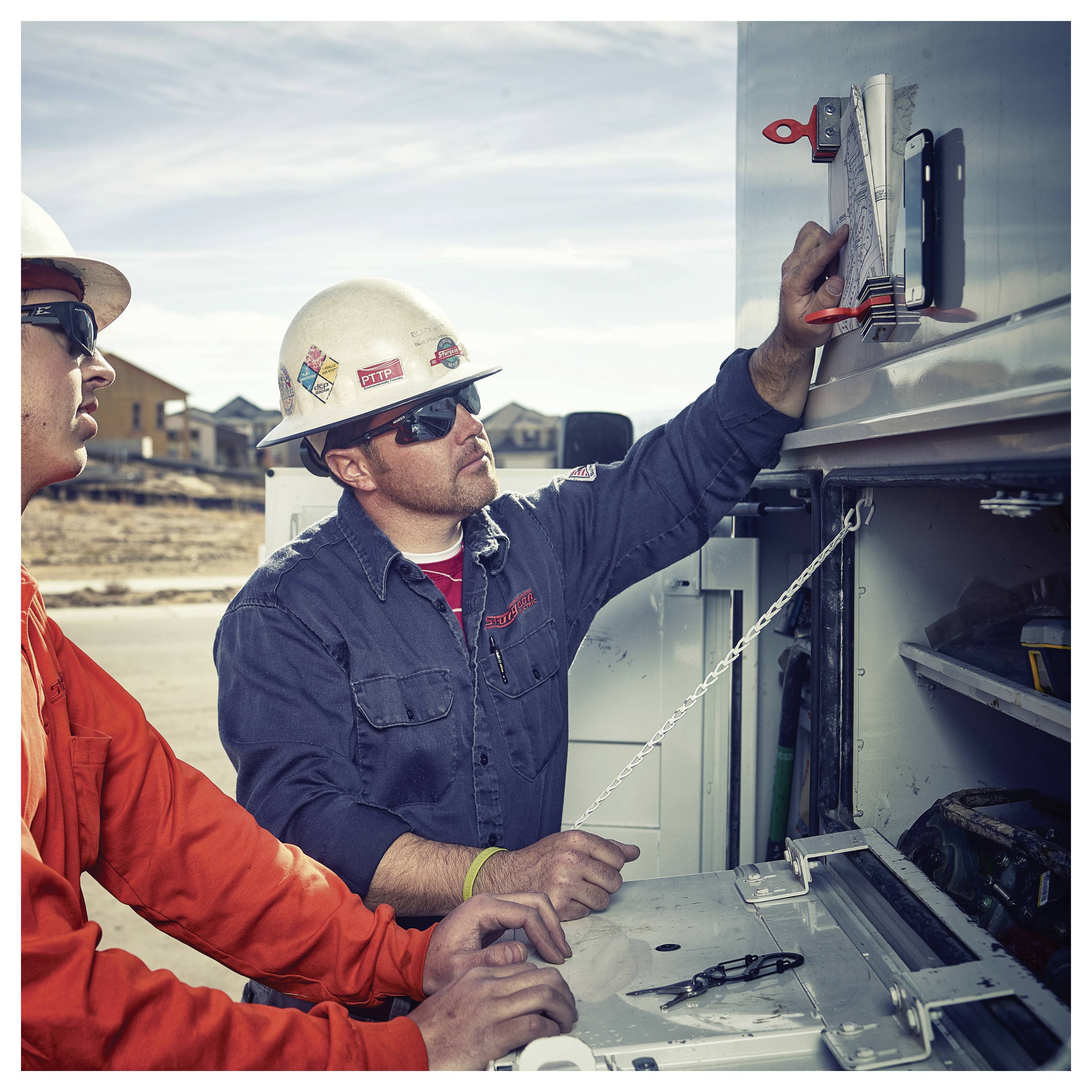 Two construction workers, one in a red jacket and the other in blue, examine equipment on a work truck at a construction site.