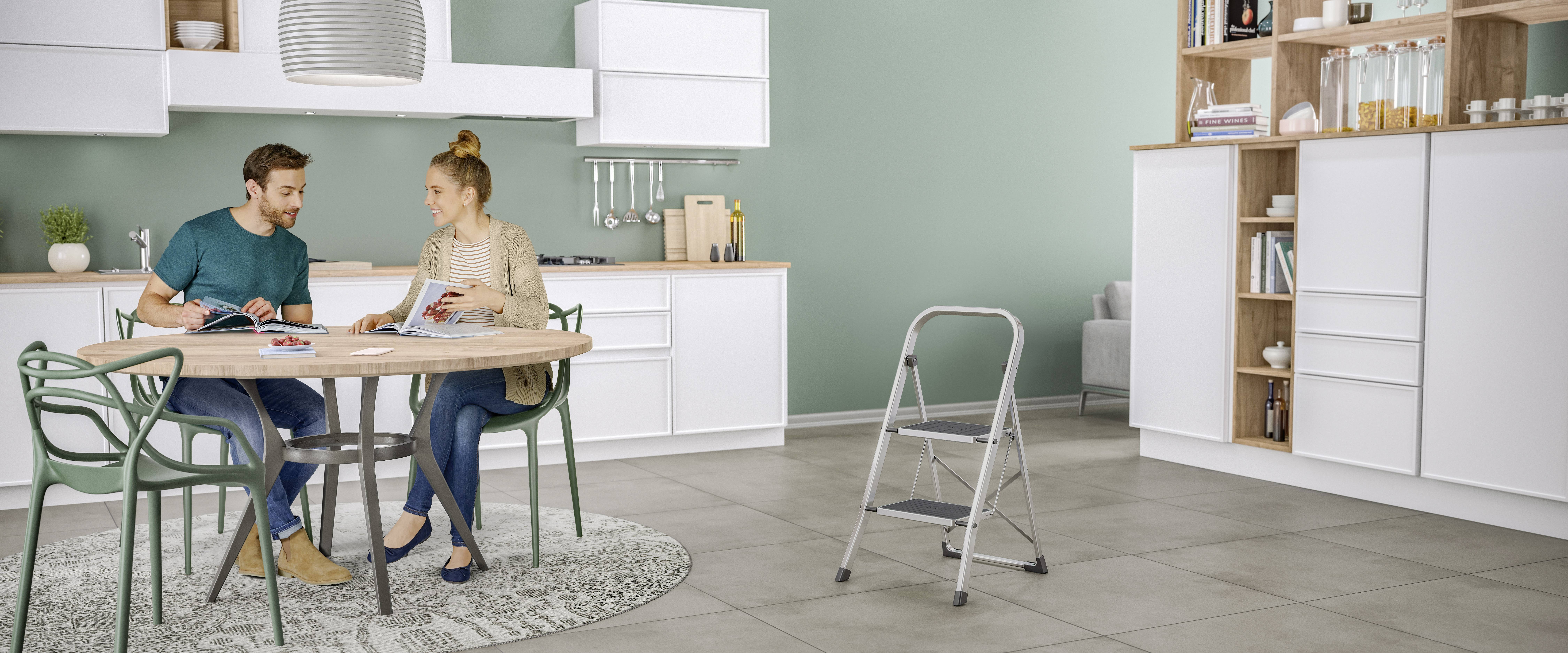 A couple sit at the kitchen table flicking through magazines in a modern kitchen with white cupboards and grey flooring.