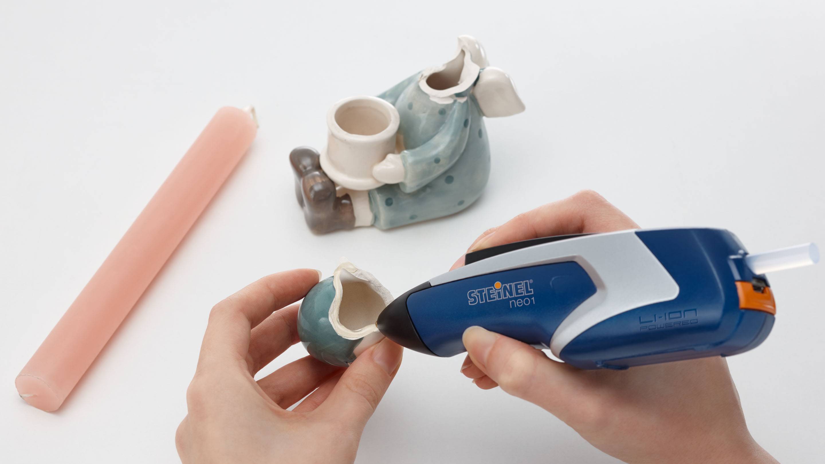 A person is repairing a broken ceramic figurine with a blue hot glue gun. A pink candle lies beside it.