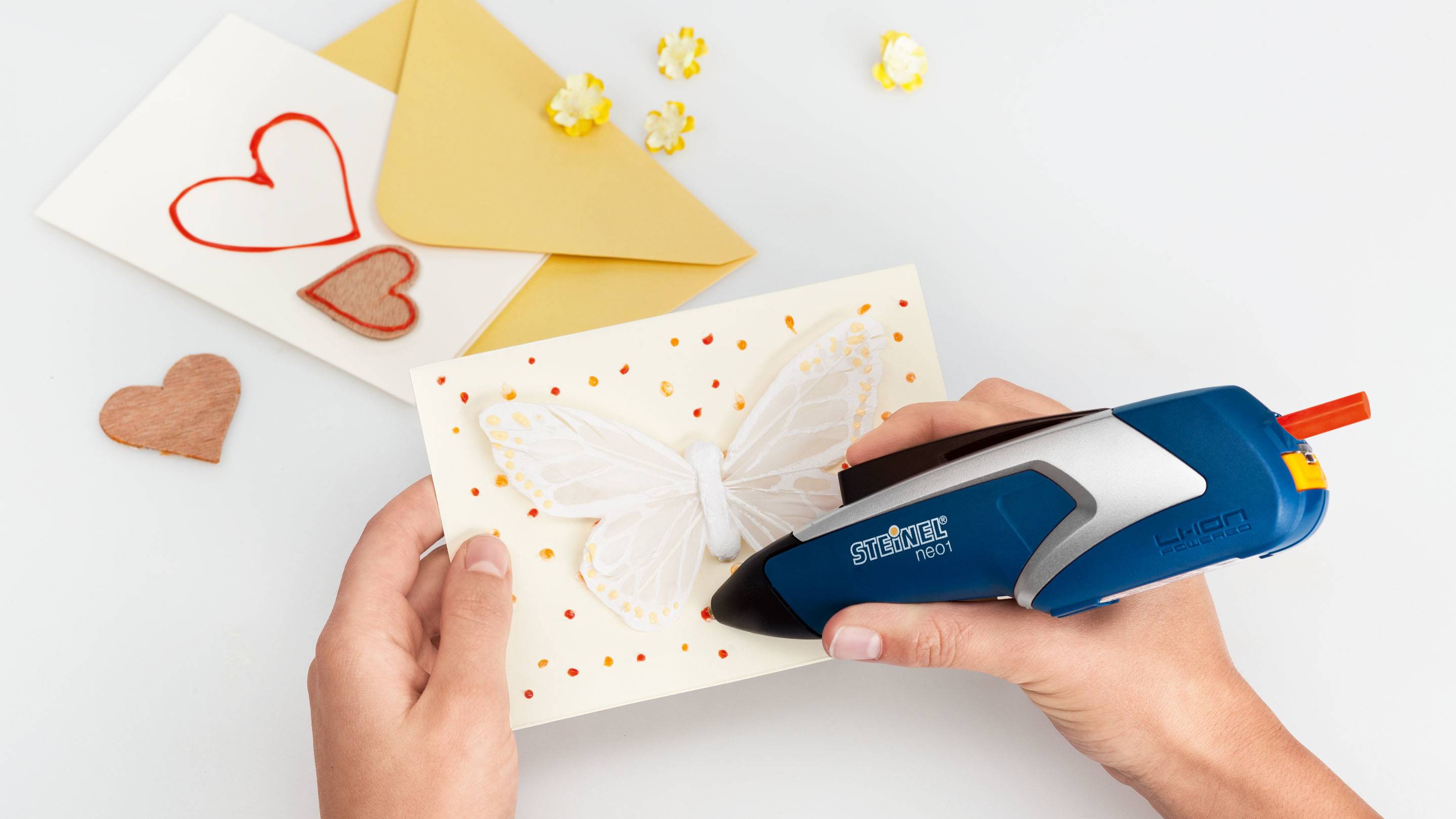 A person is decorating a card with a butterfly motif using a hot glue gun. Beside the card are an envelope and decorative hearts.