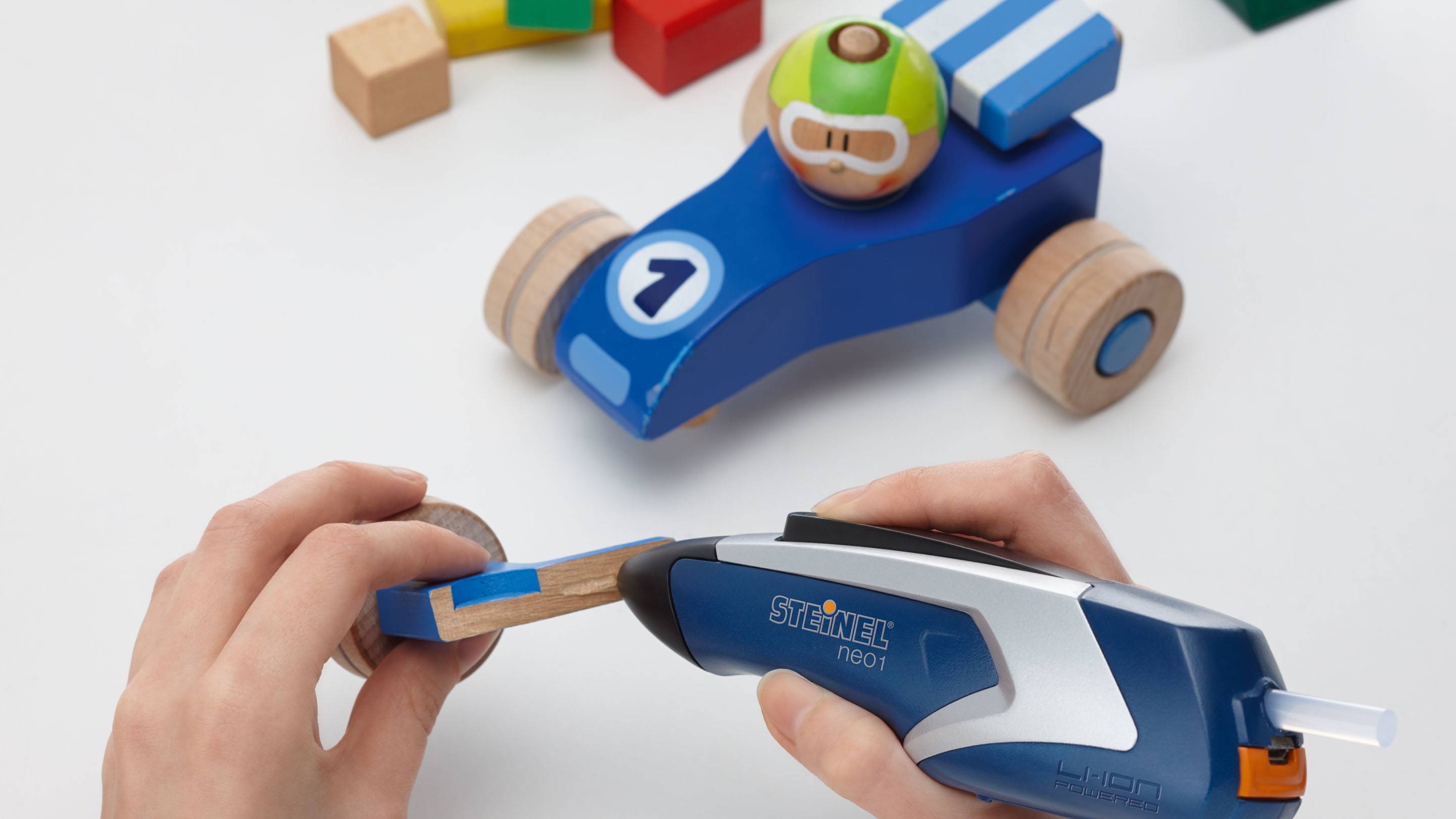 A person is attaching a wheel to a blue wooden toy car using a glue gun. Colourful wooden blocks are visible in the background.