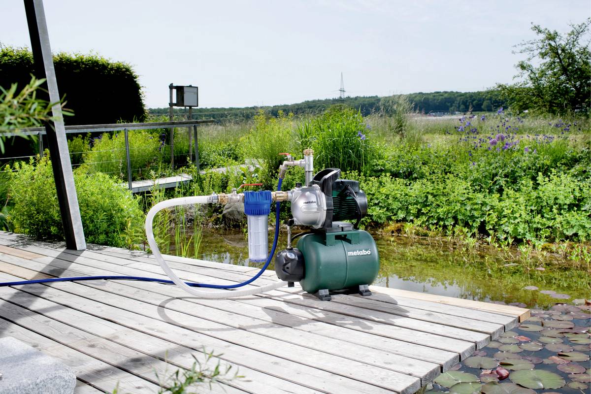 A water pump on a wooden terrace in front of a pond, surrounded by plants. In the background, a landscape with trees is visible.