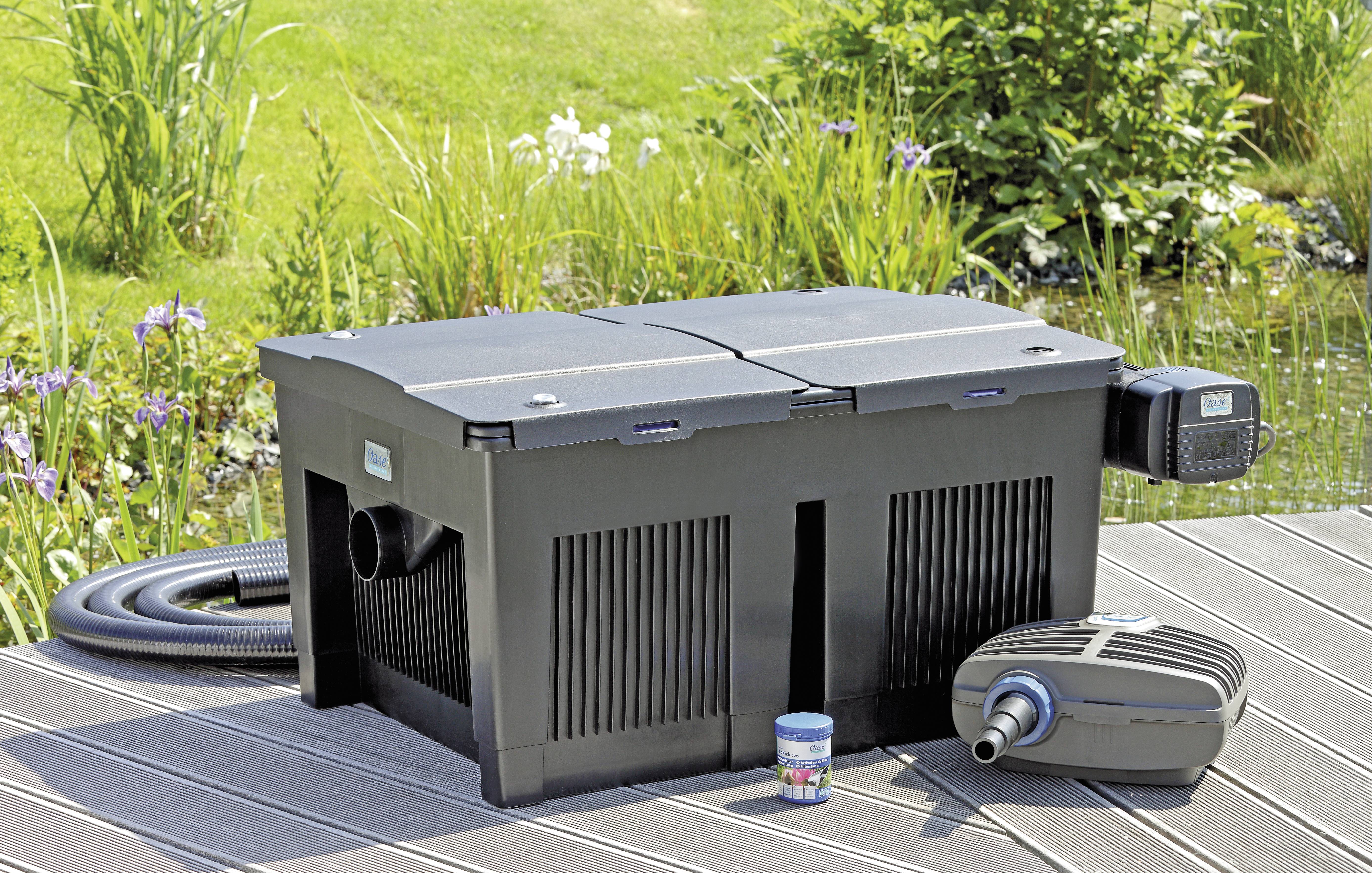 'A pond filter system displayed outdoors, set on wooden decking beside a pond. Includes a large filter box, air pump, and connecting hose.'