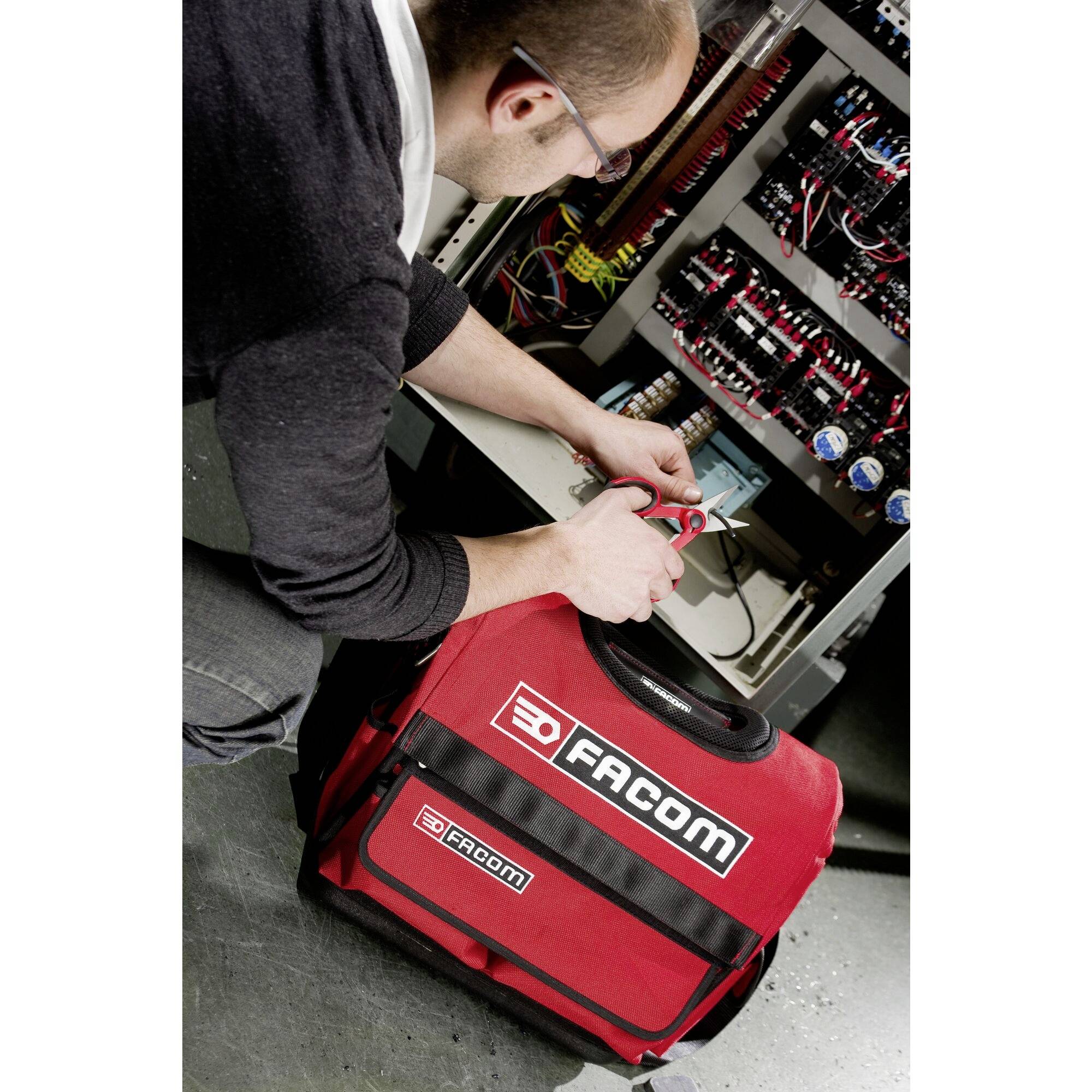 A technician is repairing an electrical installation. In front of him lies a red toolbag from the brand 'Facom'.