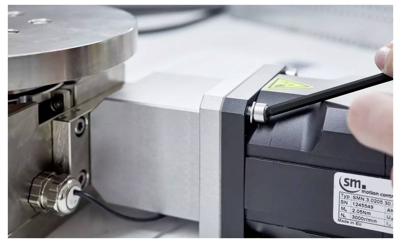 A person tightens a hex screw on a mechanical device with a label 'sm' on it. The device has metal components and electrical wiring.