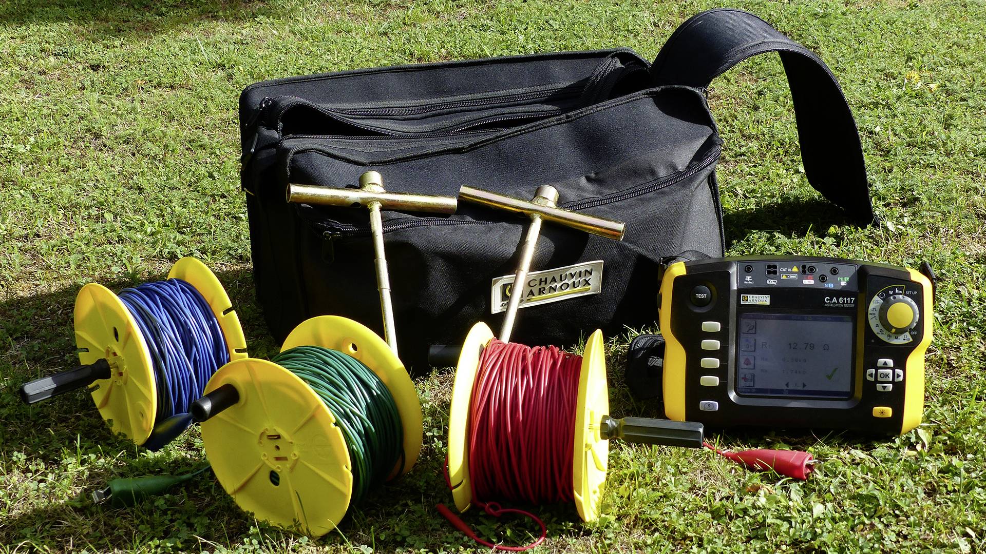 Photo of electrical testing equipment: three cable reels in yellow, blue and red, a measuring device and a black bag on grass.