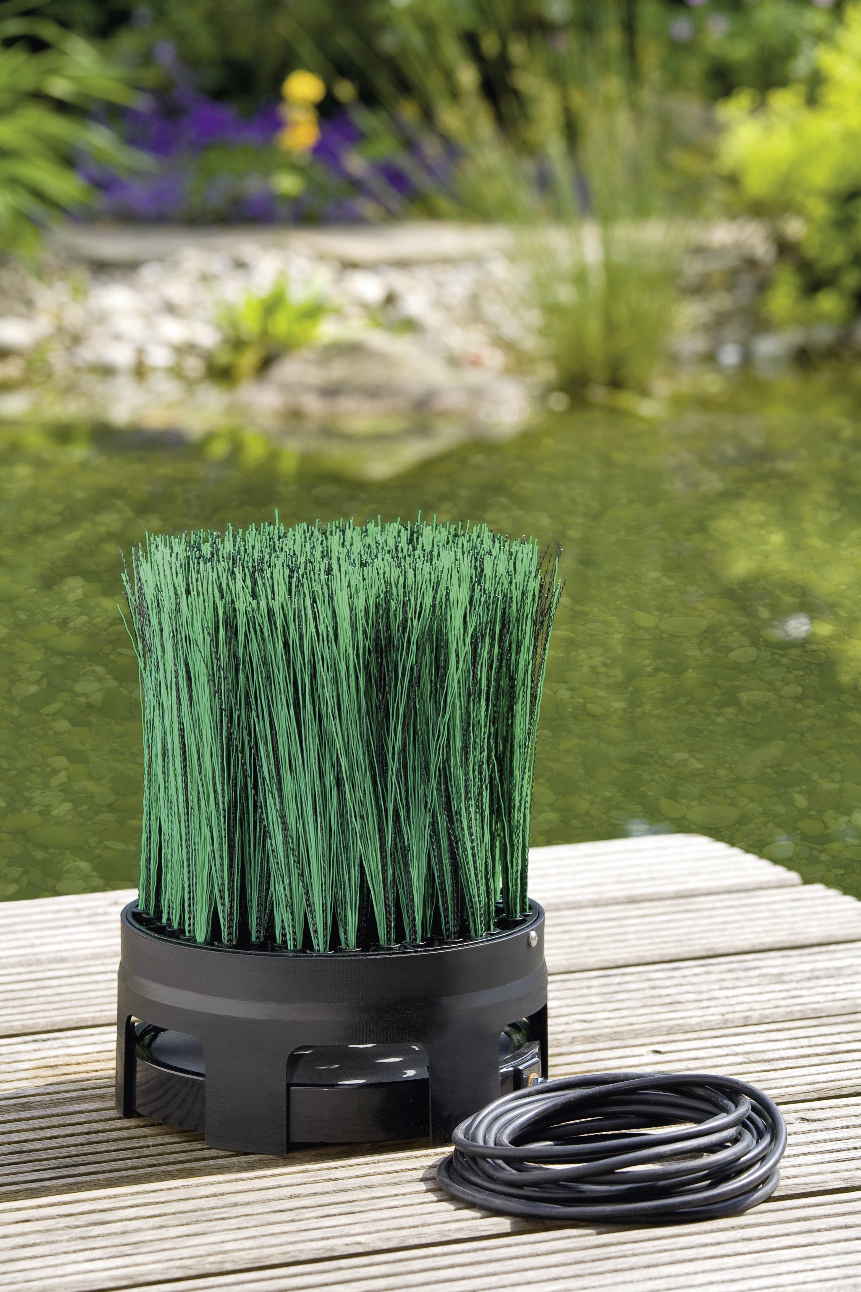 A pond filter with green bristles, placed on a wooden deck beside a coiled black cable, with water and plants in the background.