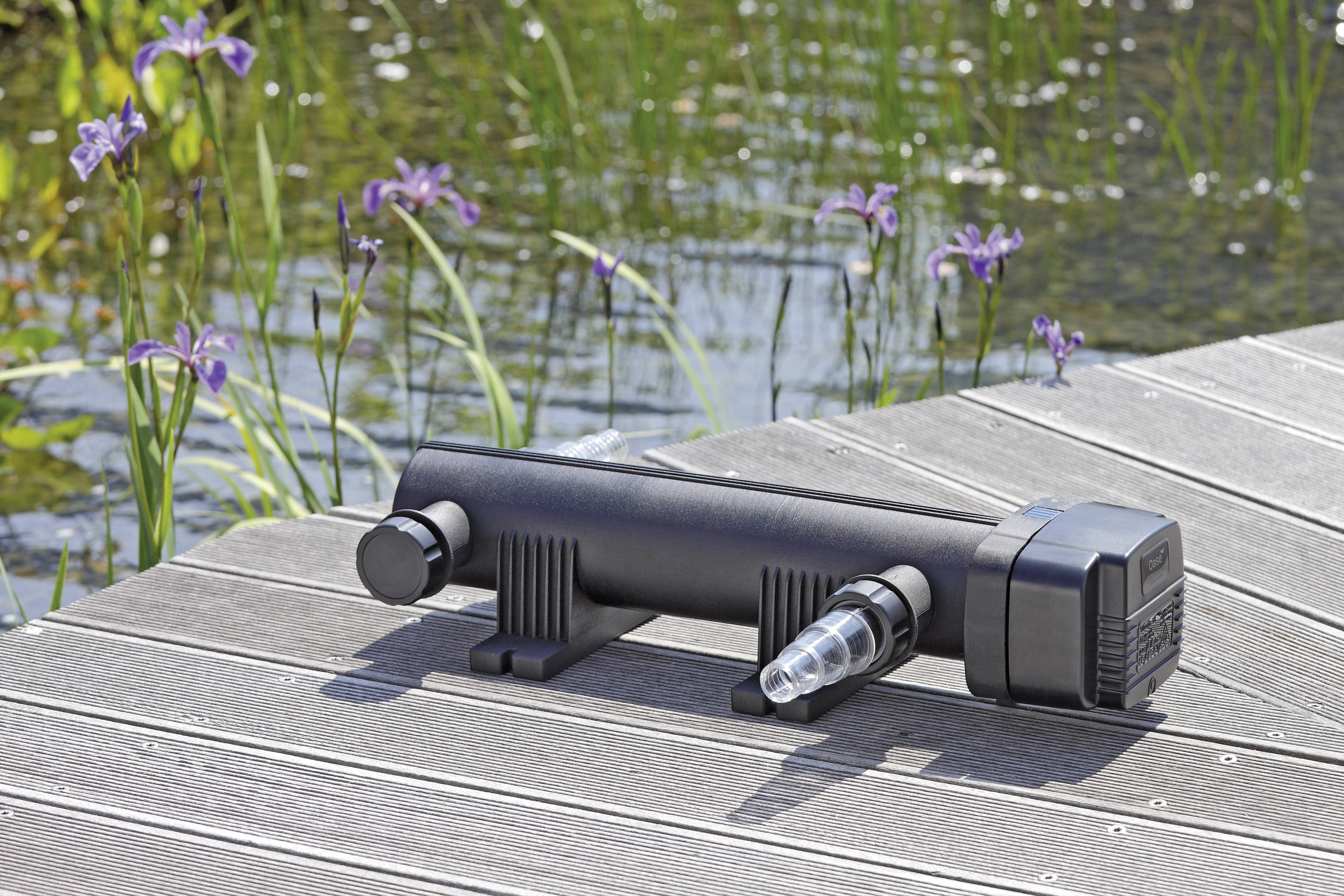 A black water pump with clear tubing is placed on a wooden deck near a pond, surrounded by purple flowers and green plants.