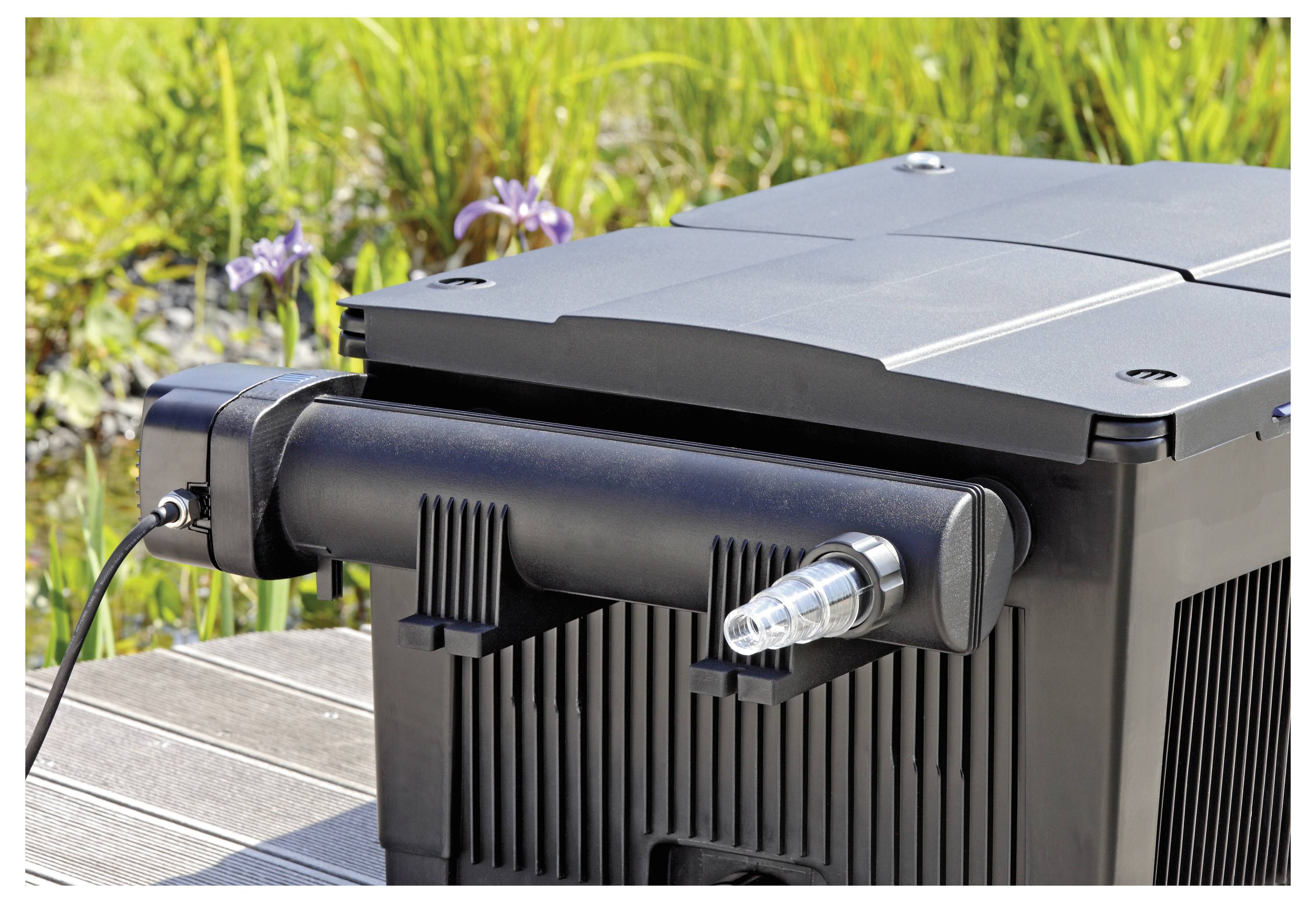 A black plastic filter box with a ribbed surface, featuring a hose attachment, is positioned outdoors on a wooden surface with greenery in the background.