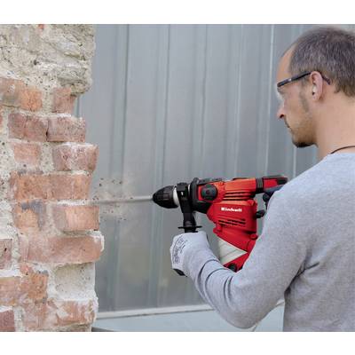 A person is drilling into a brick wall with a red drill hammer. They are wearing safety glasses and gloves.