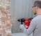 A person is drilling into a brick wall with a red drill hammer. They are wearing safety glasses and gloves.