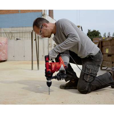 'A worker kneels on a construction site and uses a red drill on a concrete floor. The background shows construction activities.'