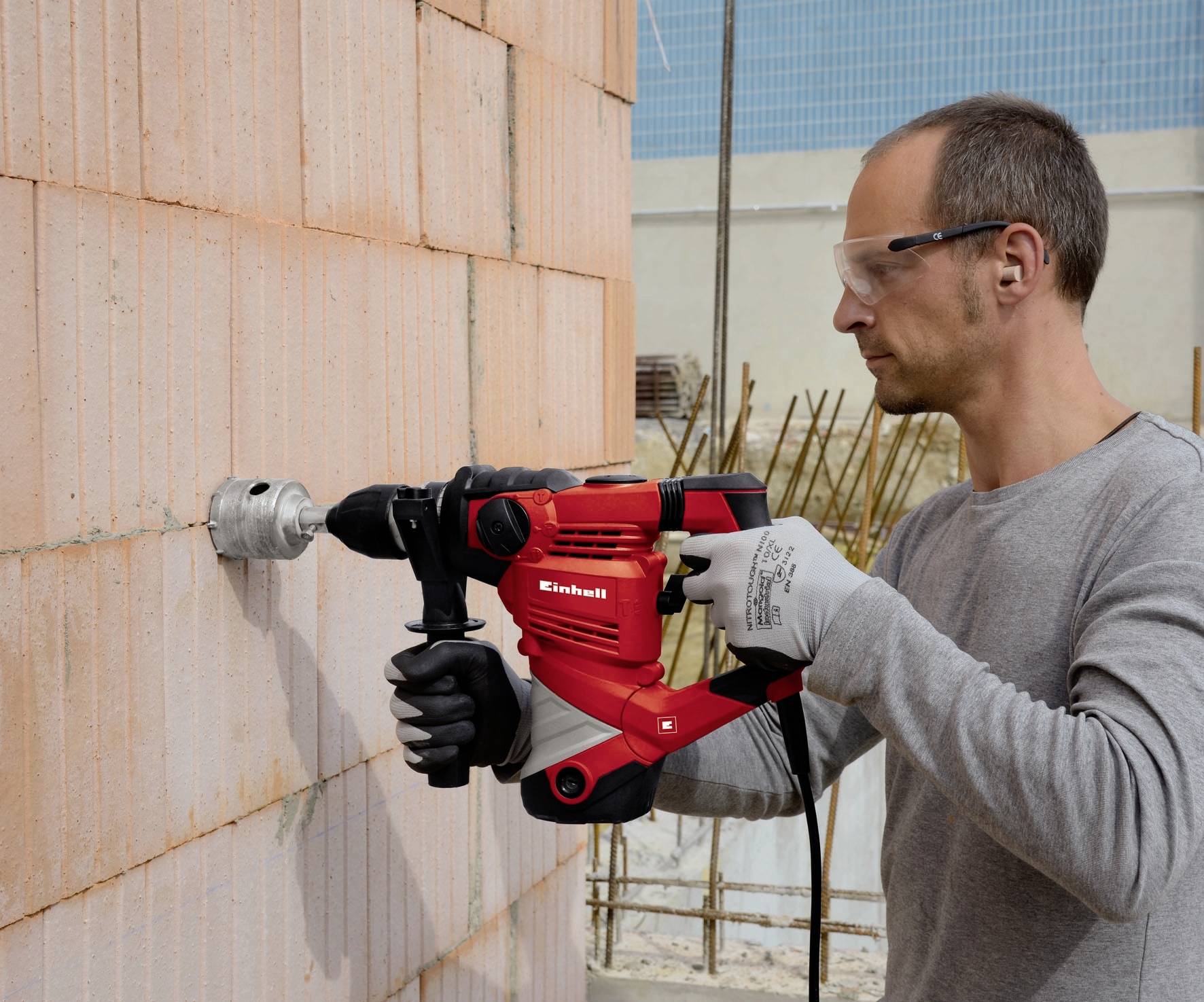 A man wearing safety glasses is using a red drill to bore a hole in a brick wall.
