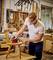 A person is sanding a wooden chair with a sanding machine in a workshop. Tools and wood can be seen in the background.
