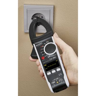 A person is holding a digital clamp multimeter in front of a wall socket to perform electrical measurements.