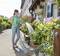 A person is watering plants along a pavement in front of a house using a garden hose. Various flowering plants are visible.