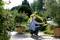 A woman waters a garden with a hosepipe on a sunny day. Surrounded by flowering plants and potted trees.