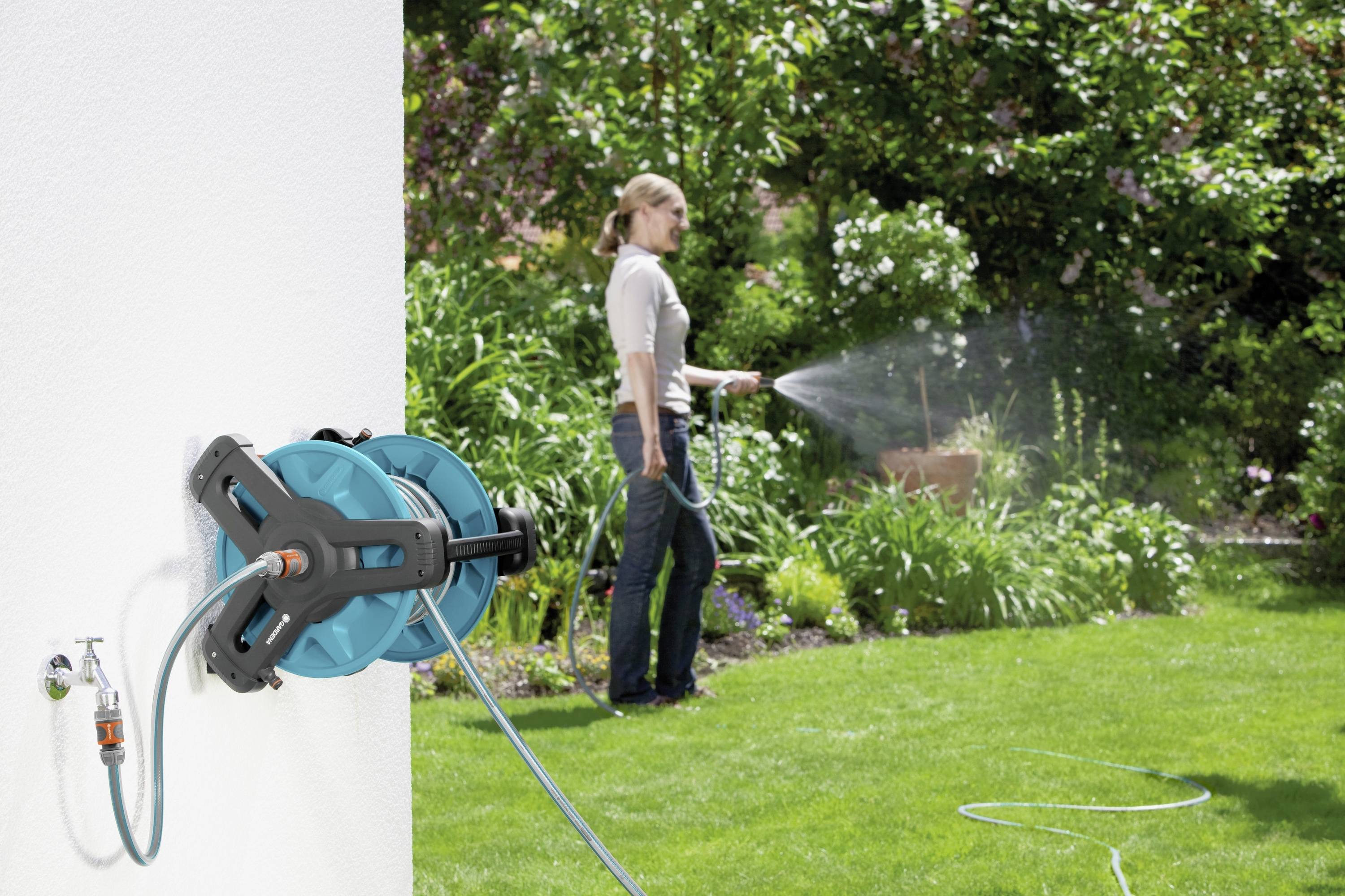 A person is watering the garden with a hose attached to a wall mount. Flowering plants are visible in the background.