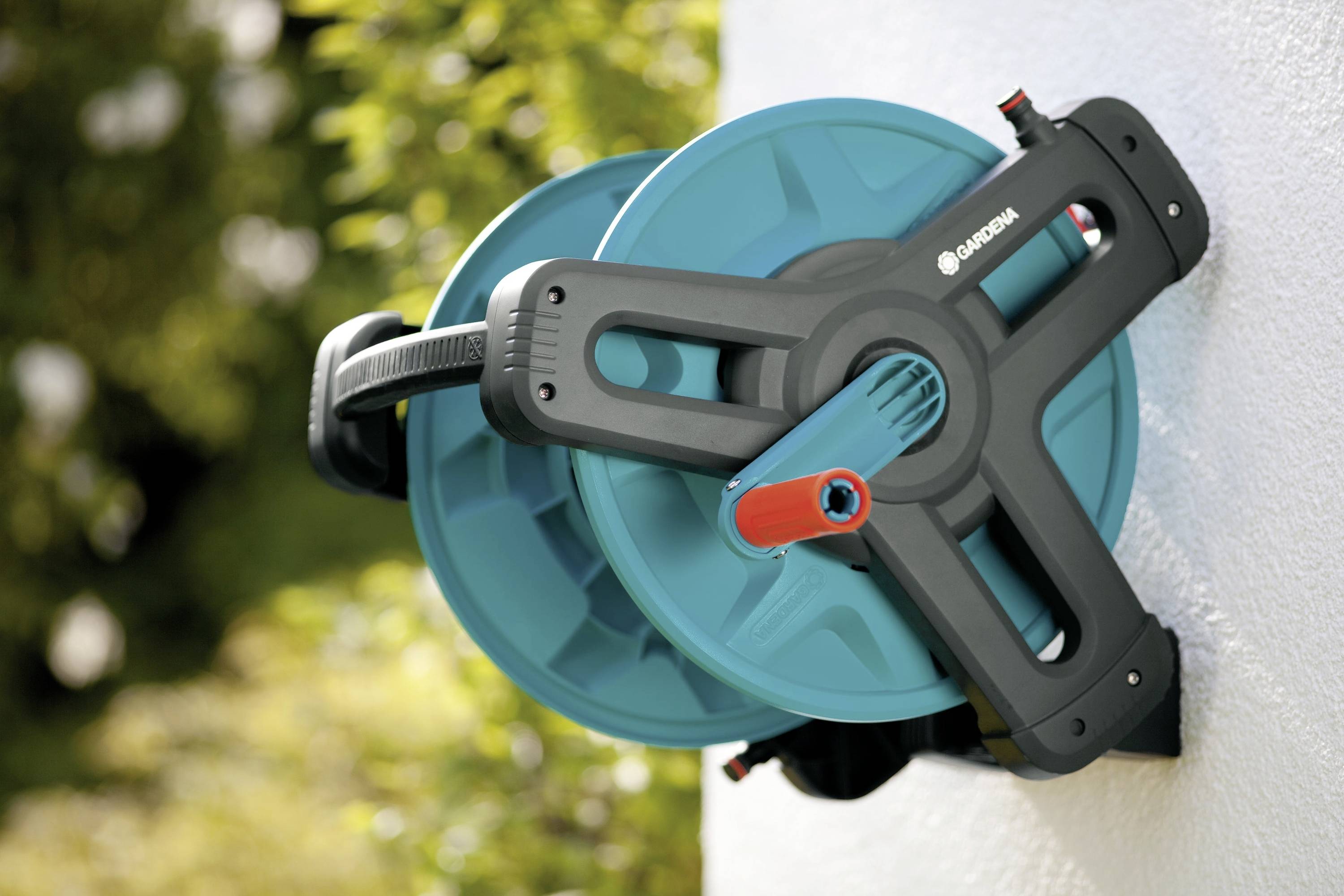 A wall-mounted hose box in blue and black, fitted to a white wall. It is surrounded by a garden with green plants.