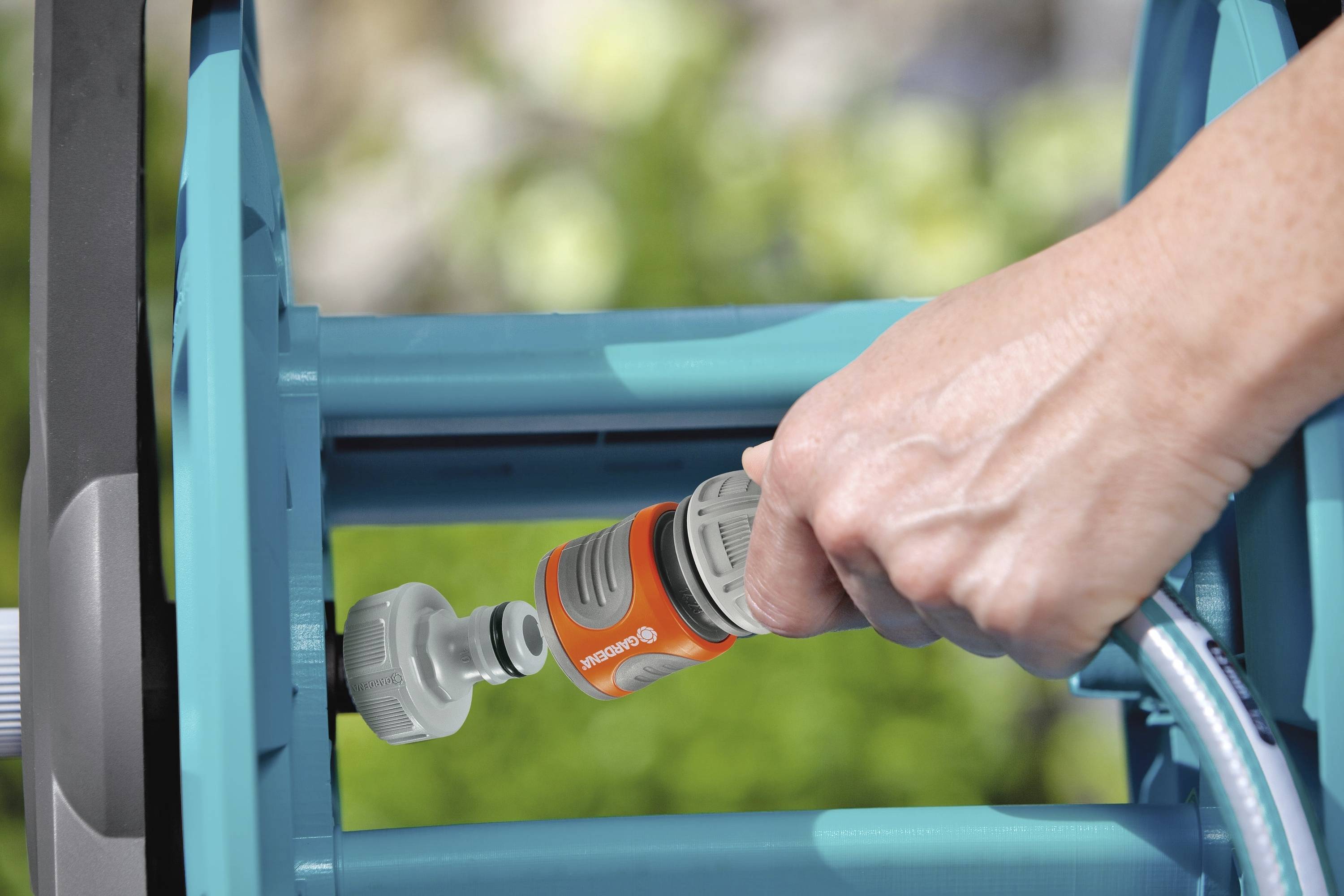 A hand holds a garden hose and connects it to an orange hose connector on a blue hose reel.