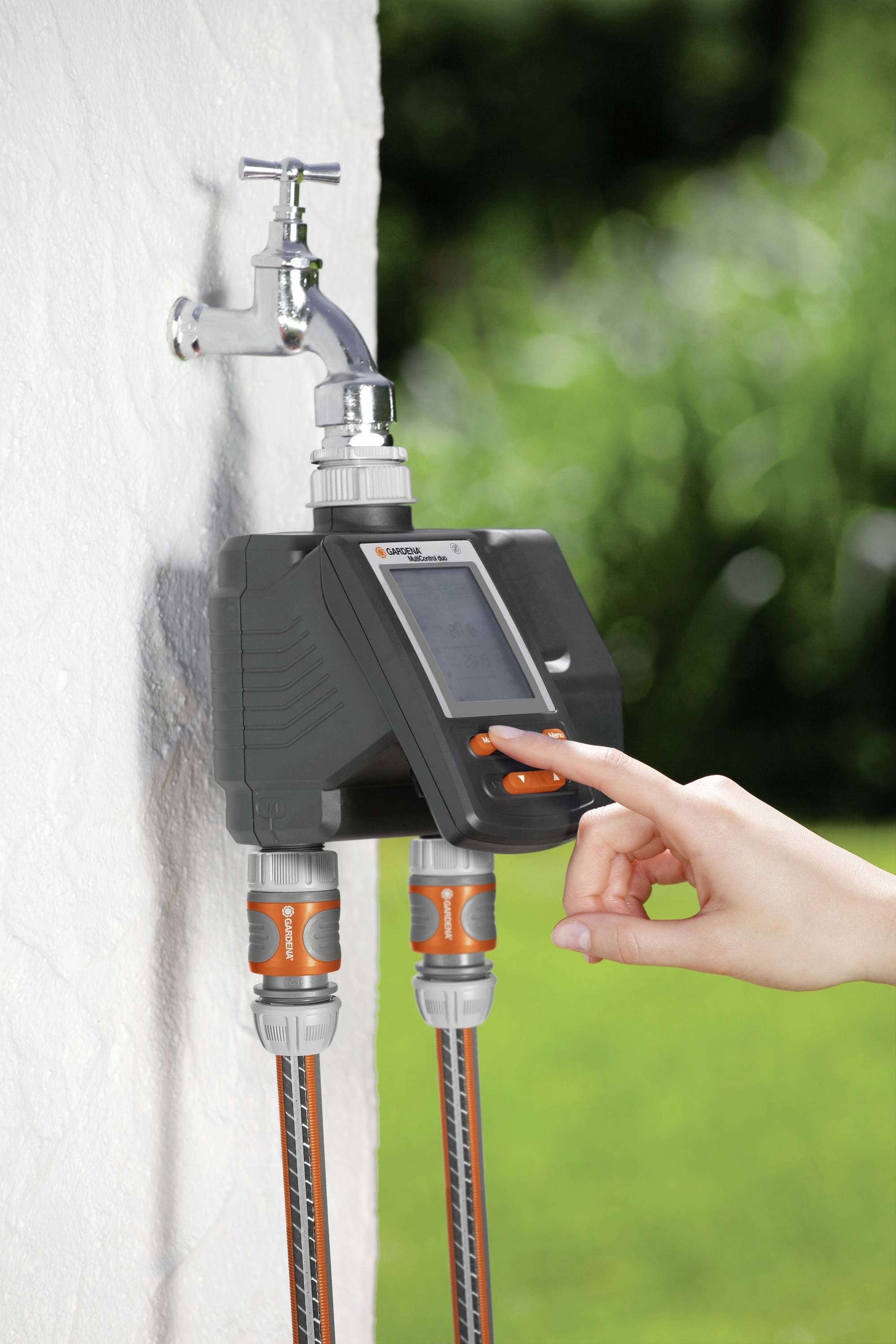 A hand operates a digital irrigation device attached to an outdoor tap. Two hoses are connected.