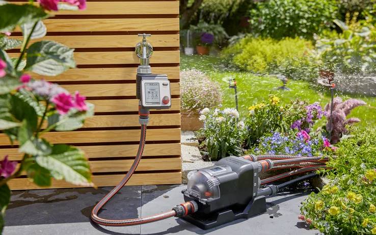 A garden irrigation system automatically watering plants with a hose connected to an outdoor wall, surrounded by flowers and greenery.