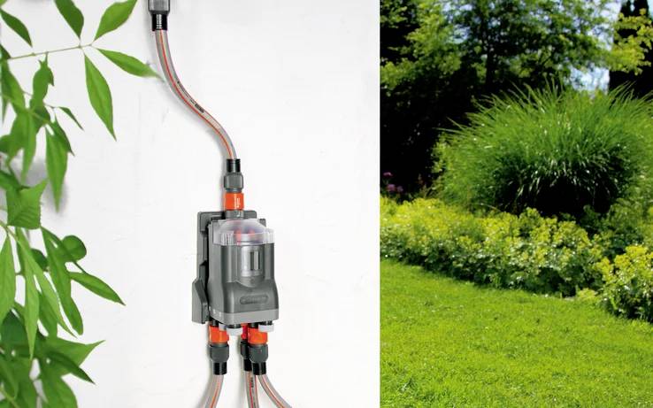'An irrigation system control unit mounted on a white wall. Red and black hoses extend from it, leading to a lush green garden.'