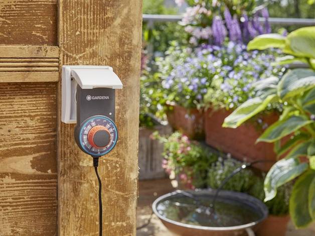 An automatic garden irrigation system is mounted on a wooden fence. In the background, flowering plants and a small fountain can be seen.