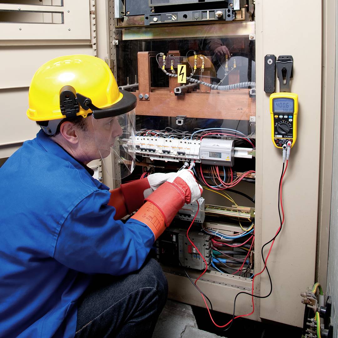 An electrician in blue workwear and a yellow safety helmet is working on an open electrical cabinet. Instruments and cables are visible.