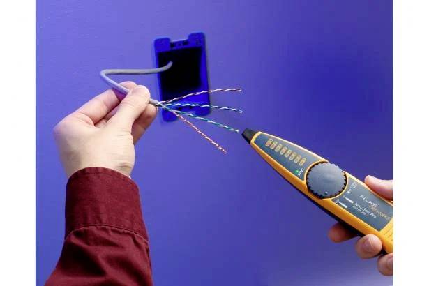 A person is testing cables with a cable testing device in front of a blue wall. The cables are colourfully twisted and emerge from a wall opening.
