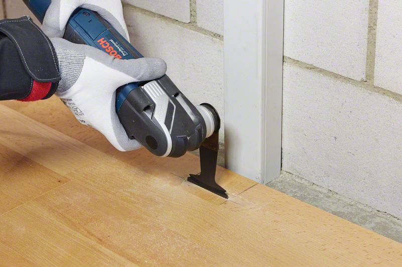 A person is using a multi-tool to cut a wooden floor precisely at the edge. They are wearing protective gloves.