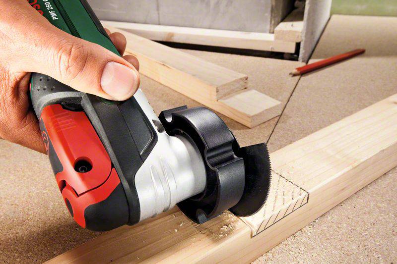 A hand is holding an electric multi-tool cutting a rectangular pattern into a piece of wood.